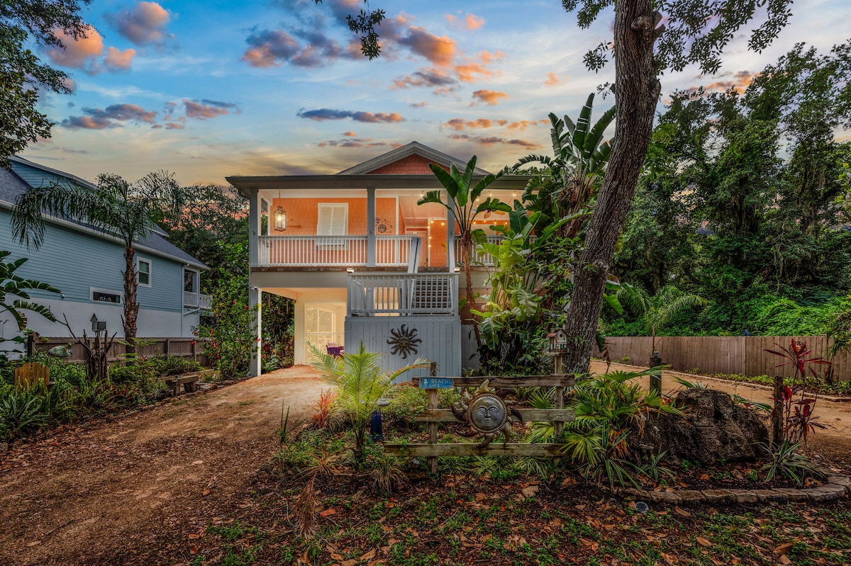 The house is elevated with a staircase leading to the front entrance. Lush greenery and tropical plants surround the property, while a gravel driveway provides access. The sky is colorful at dusk, enhancing the charming presence of the home.