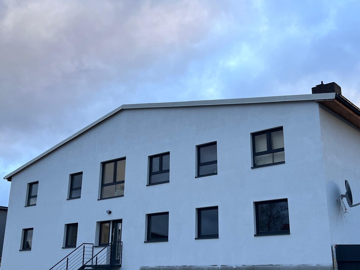 The exterior of a two-story building is shown, featuring a white facade and multiple windows. The entryway is accessible with a set of stairs leading to the front door. A satellite dish is mounted on the side, indicating connectivity options.