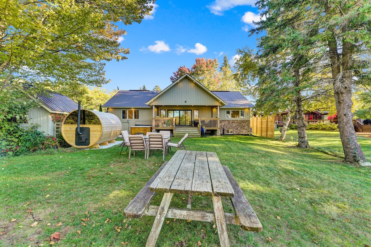 The serene outdoor space features a large grassy area with a wooden picnic table and several lounge chairs. A wood-burning barrel sauna is positioned near the house, surrounded by well-maintained landscaping and trees. The home's exterior reflects a modern design with large windows and a welcoming facade.