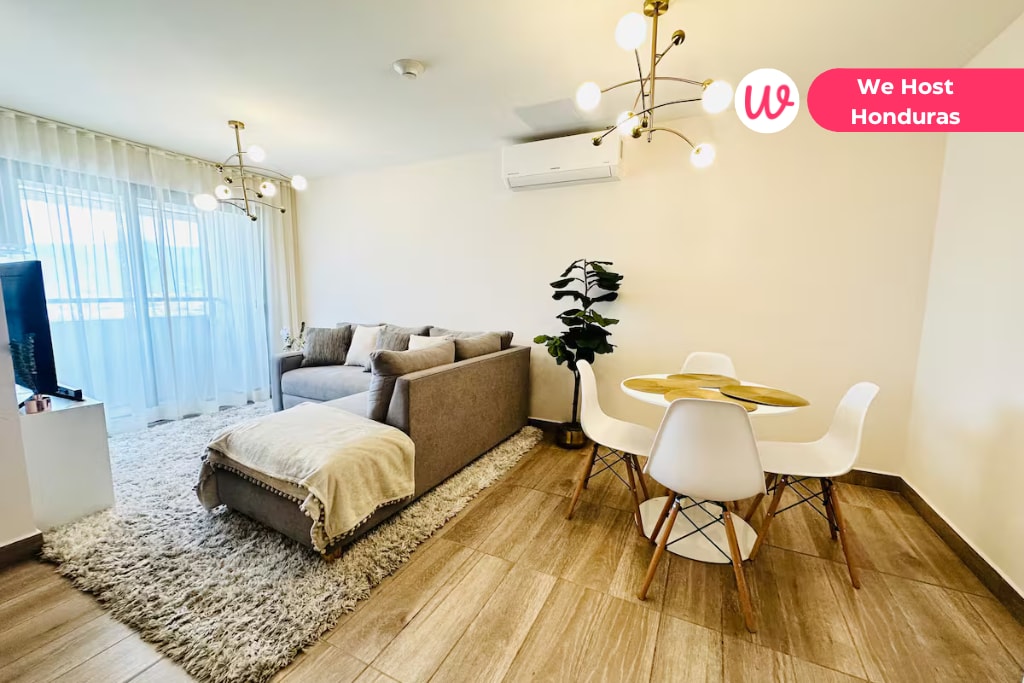 A bright living space features a comfortable gray sectional sofa paired with a cozy throw blanket. A modern dining table with white chairs sits nearby. Natural light filters through sheer curtains, highlighting the warm tones of the wood-look flooring.