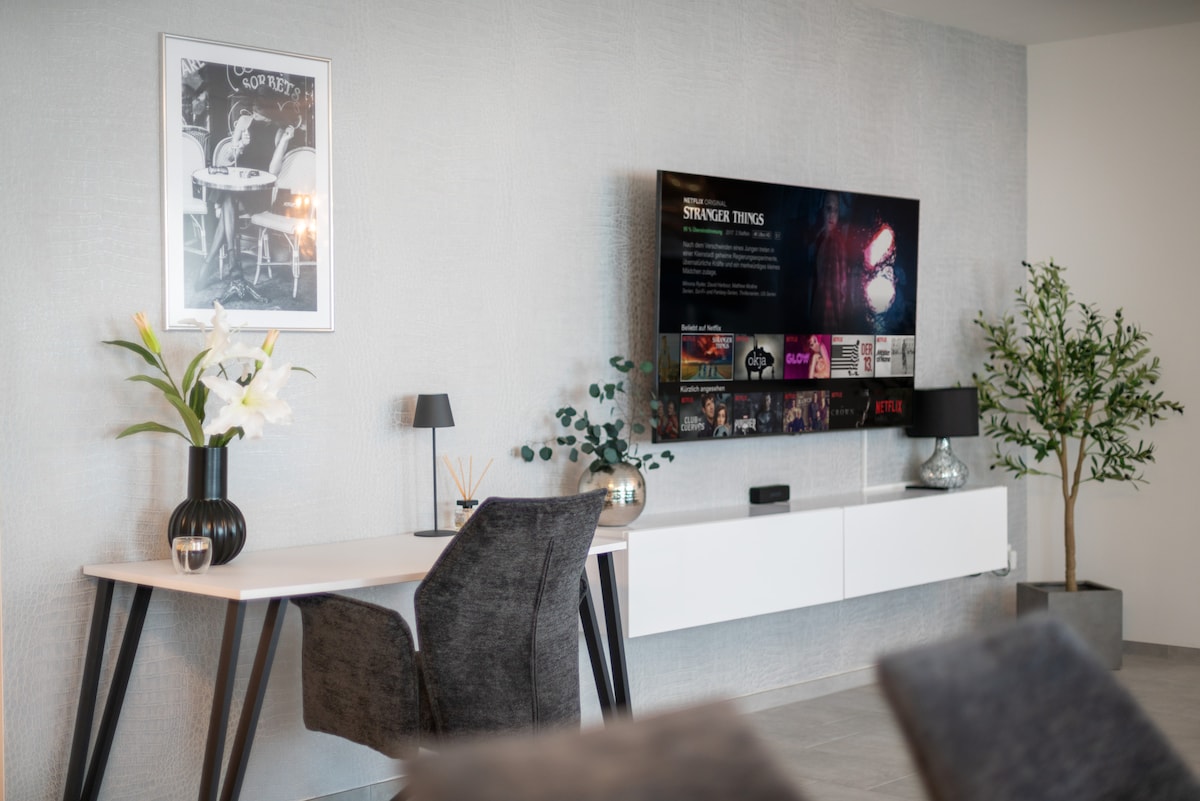 An inviting living space is depicted, featuring a wall-mounted TV displaying a streaming service. A sleek white desk with a modern chair is positioned nearby, accompanied by decorative plants and a stylish lamp. Fresh flowers add a touch of elegance to the decor.