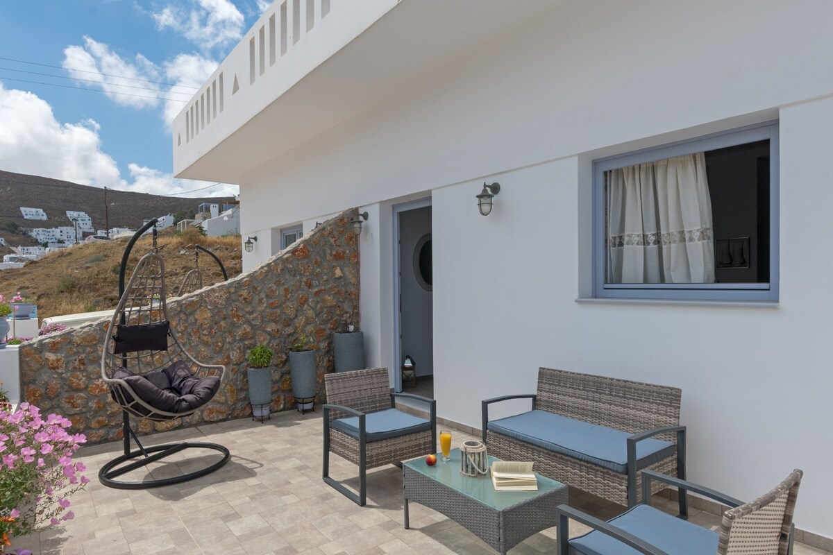 An outdoor terrace area features modern woven furniture, including a seating set and a round hanging chair. Potted plants add greenery, while a small table displays a drink. The surrounding stone wall enhances privacy, and the scenery offers a glimpse of the hillside.