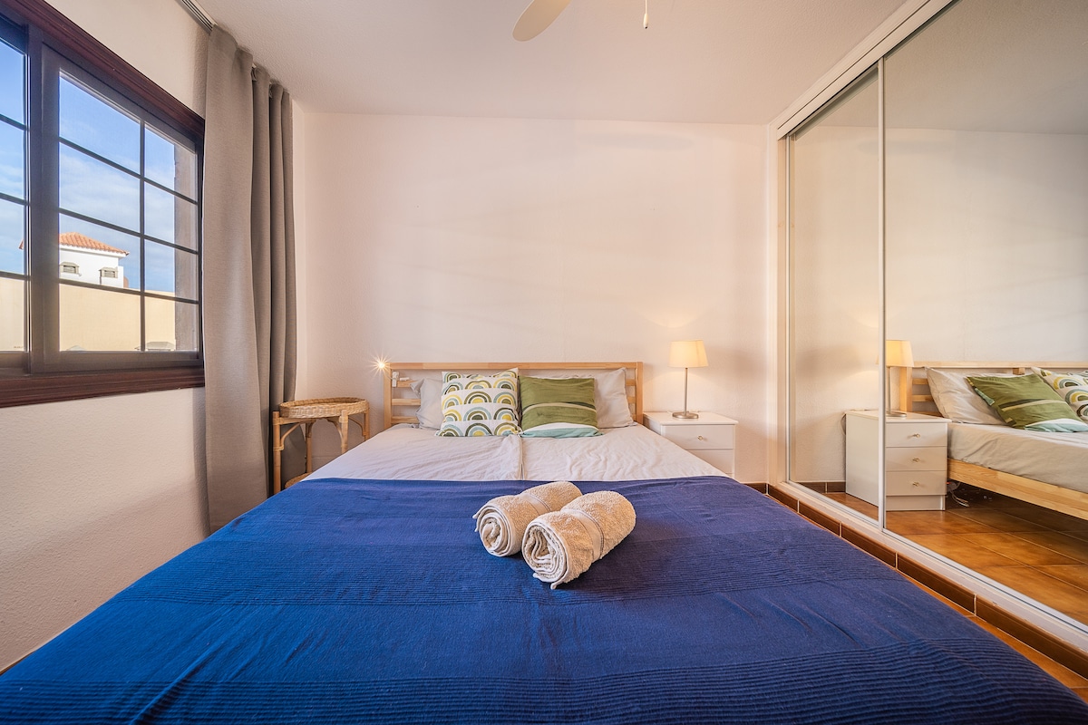 A modern bedroom features a spacious bed with crisp linens and decorative pillows. A bedside lamp provides soft lighting beside the bed, while a large mirror enhances the sense of space. Natural light filters through the window covered by light drapes.