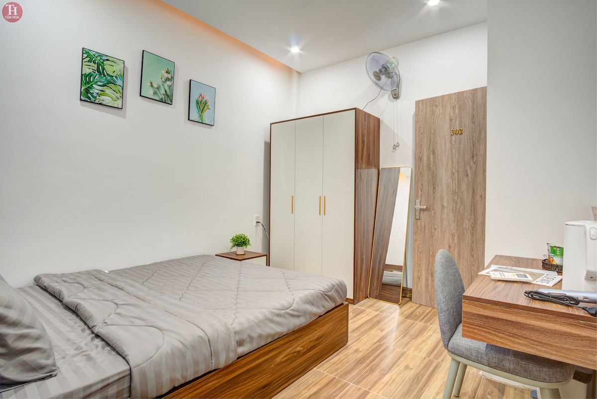 A well-lit bedroom features a spacious bed with a neutral-toned quilt. Simple artwork adorns the wall above, while a dual-door wardrobe provides storage. A small table and chair are positioned near a bright window, creating a functional work area.