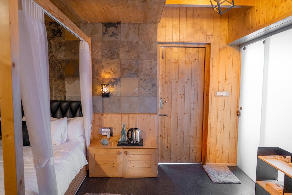 A cozy couple's retreat room features a spacious canopy bed with soft drapery, complemented by wooden accents on the walls. A minimalist wooden cabinet and kettle are positioned nearby, while light brightens the space through the open door, creating a warm and inviting atmosphere.