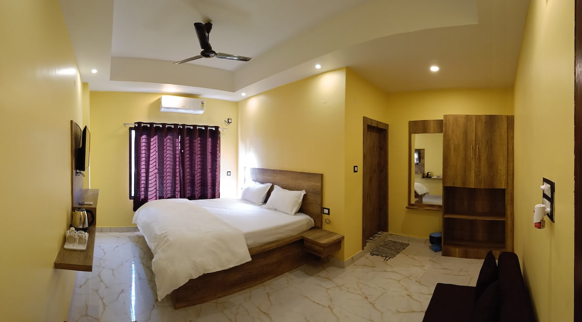 A bright bedroom features a spacious bed with crisp white linens against a warm yellow wall. Dark curtains frame a window, and a wooden dresser is visible beside a mirror. A seating area with a brown sofa is positioned in the corner, adding a cozy touch.