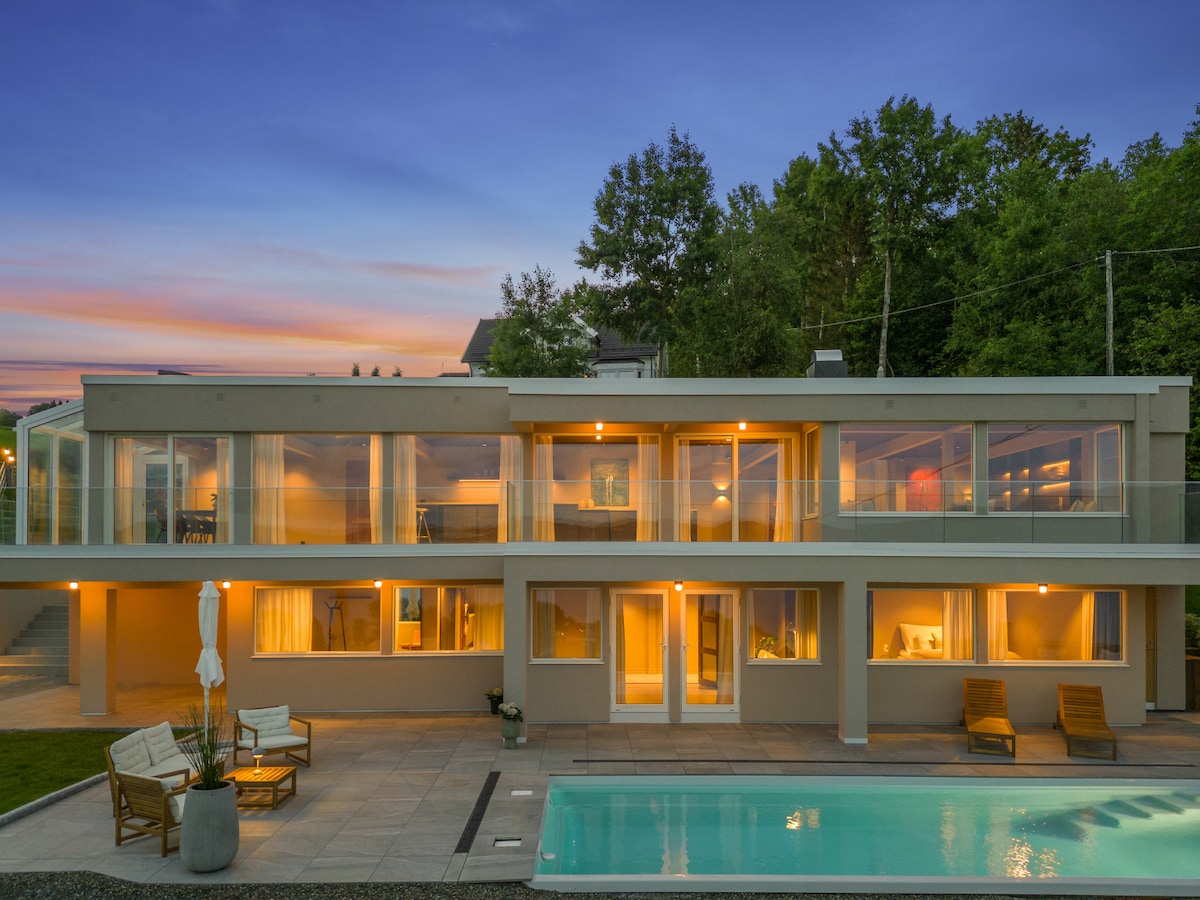 The villa's exterior features a modern design, illuminated by warm evening light. A spacious outdoor area includes a calm swimming pool surrounded by lounge chairs, providing a serene atmosphere. Expansive glass panels create a connection between the indoor and outdoor spaces.