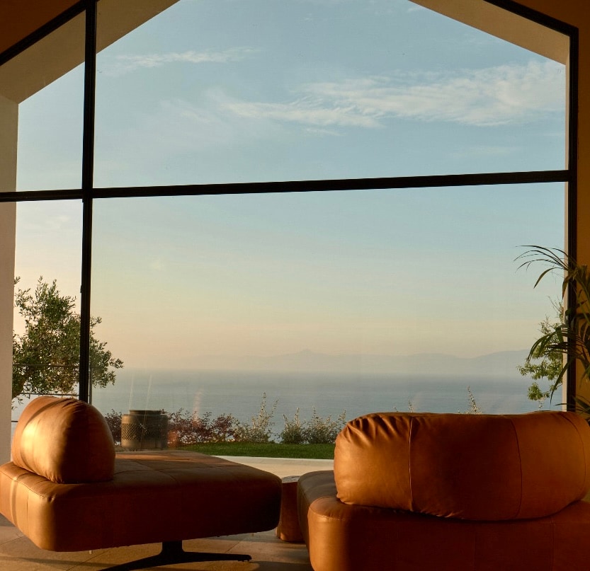 A spacious seating area features modern, curved leather sofas positioned before large glass windows, offering unobstructed views of the sea and distant horizon. Soft natural light enhances the serene ambiance, while greenery is visible outside the window.
