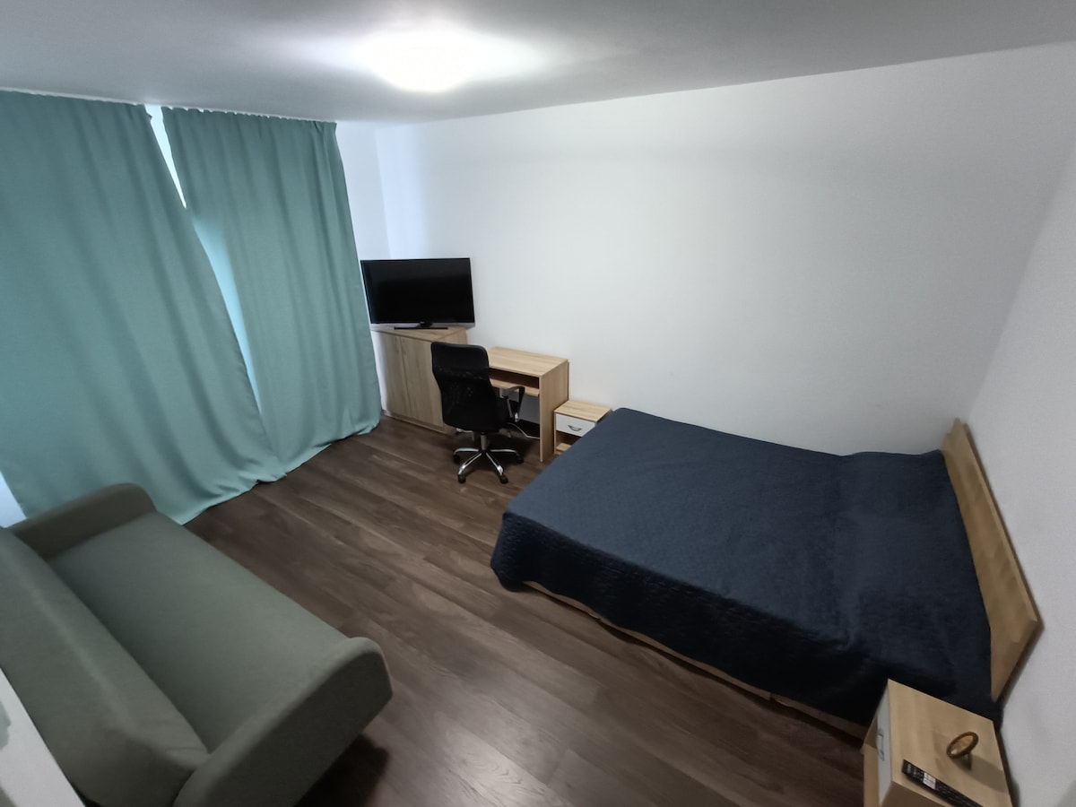 A clean and modern room features a spacious bed covered with a dark blue blanket. Light wood furniture includes a desk with a chair and a bedside table. Large, light teal curtains frame a window, along with a television mounted on the wall.
