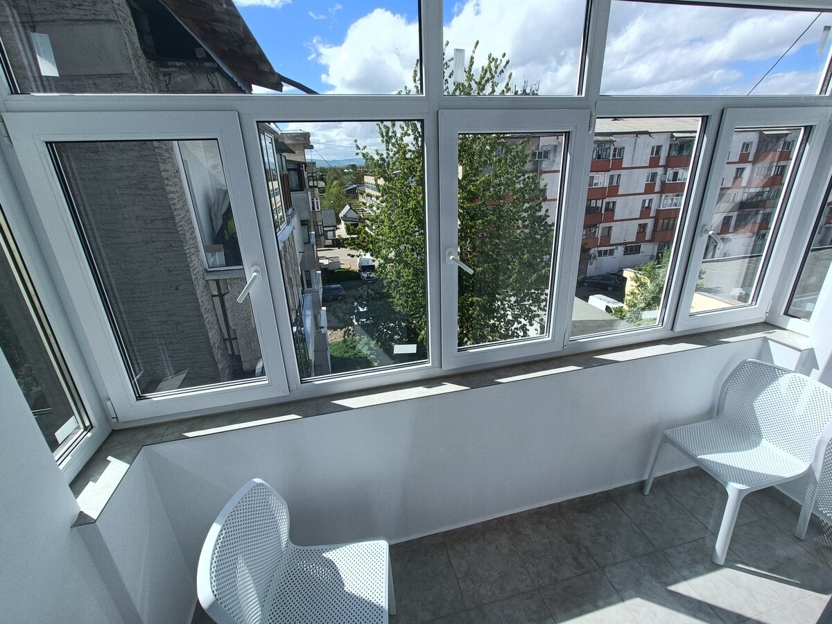 A bright sunroom features multiple large windows that provide a panoramic view of the surrounding area, including greenery and neighboring buildings. Two lightweight chairs are positioned in the corner, creating a cozy spot for relaxation.