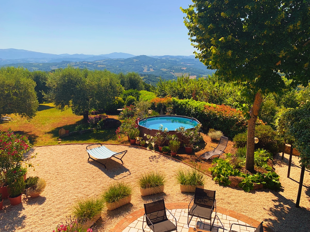 A seasonal pool is surrounded by well-kept gardens featuring a variety of plants, including olive trees and colorful flowers. Lounge chairs and seating areas provide spaces for relaxation, while the distant hills offer a picturesque backdrop under clear blue skies.