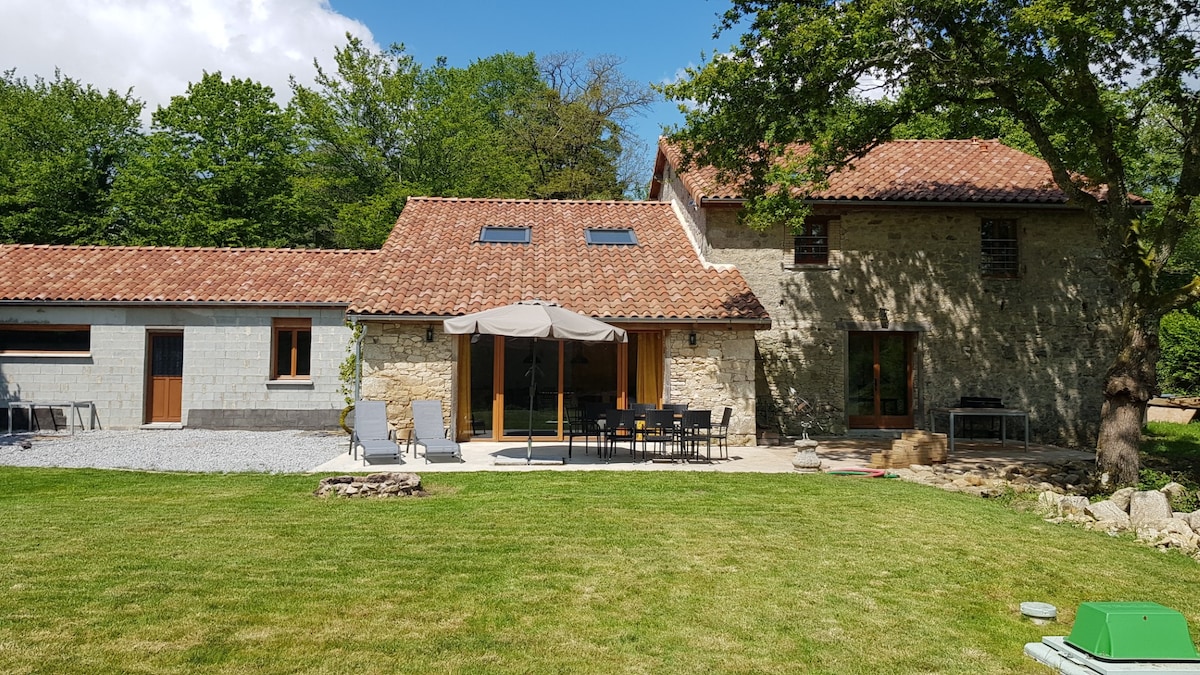 A spacious outdoor area features a well-maintained lawn with shaded seating under an umbrella. The stone exterior of the house complements the natural surroundings, with large glass doors opening to a terrace. Lush greenery frames the property, enhancing the peaceful ambiance.