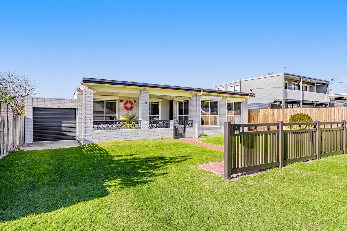 A single-story home is shown with a neatly landscaped front yard featuring green grass. The house has a light-colored exterior complemented by a red wreath on the front door. A driveway leads to a garage, and a fence encloses the property, enhancing privacy.
