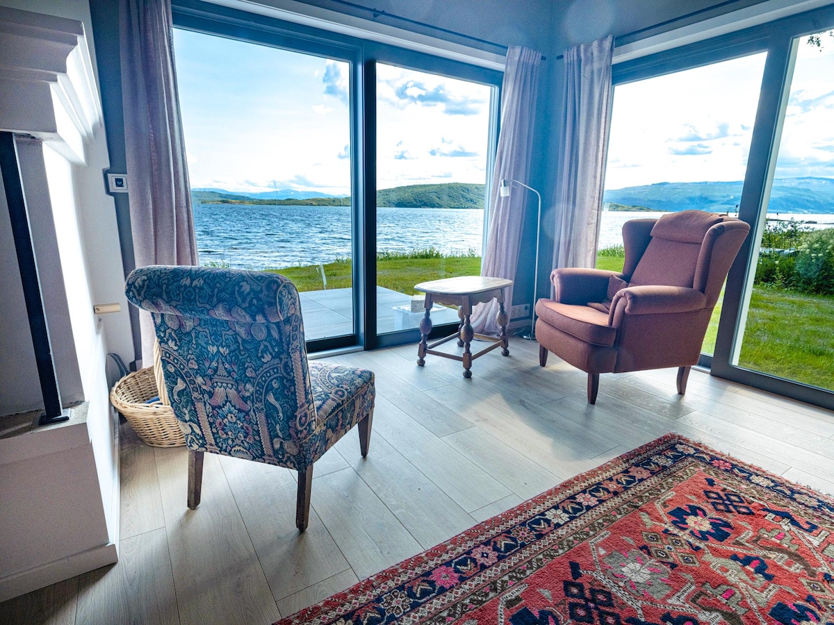 A light-filled seating area features two chairs, one patterned in blue and white and the other in a warm brown hue. A small wooden table sits between them, with expansive glass windows offering scenic views of the water and surrounding landscape.