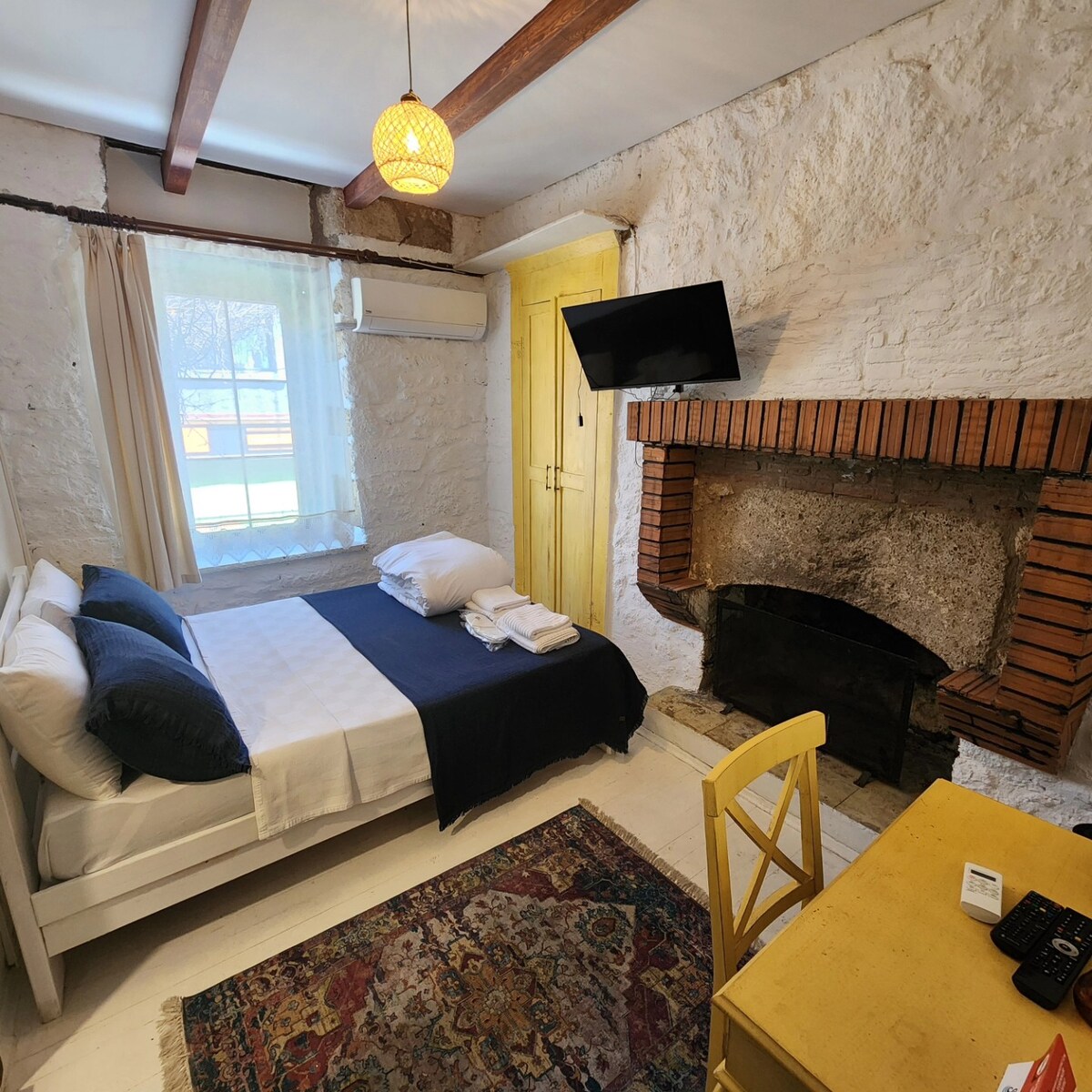 A cozy room is showcased with a spacious bed dressed in crisp linens and a navy blue throw. A rustic brick fireplace is visible, alongside an antique-style desk and chair. A window allows natural light to fill the space, complemented by a patterned area rug.