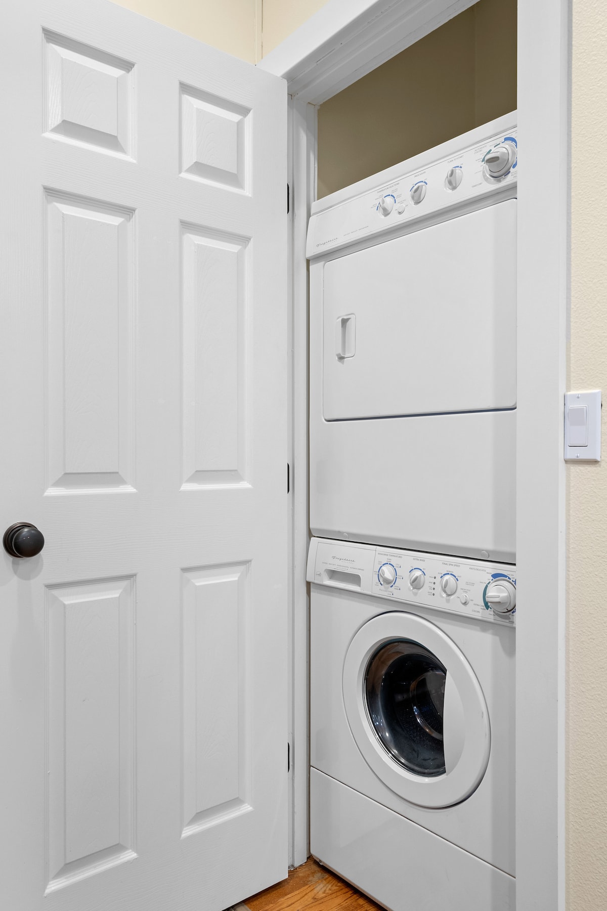 A compact laundry closet is shown, featuring a stacked washer and dryer unit. The white appliances are neatly placed within a light-colored wall enclosure, with a closed door that maintains a tidy appearance. Wooden flooring complements the space.
