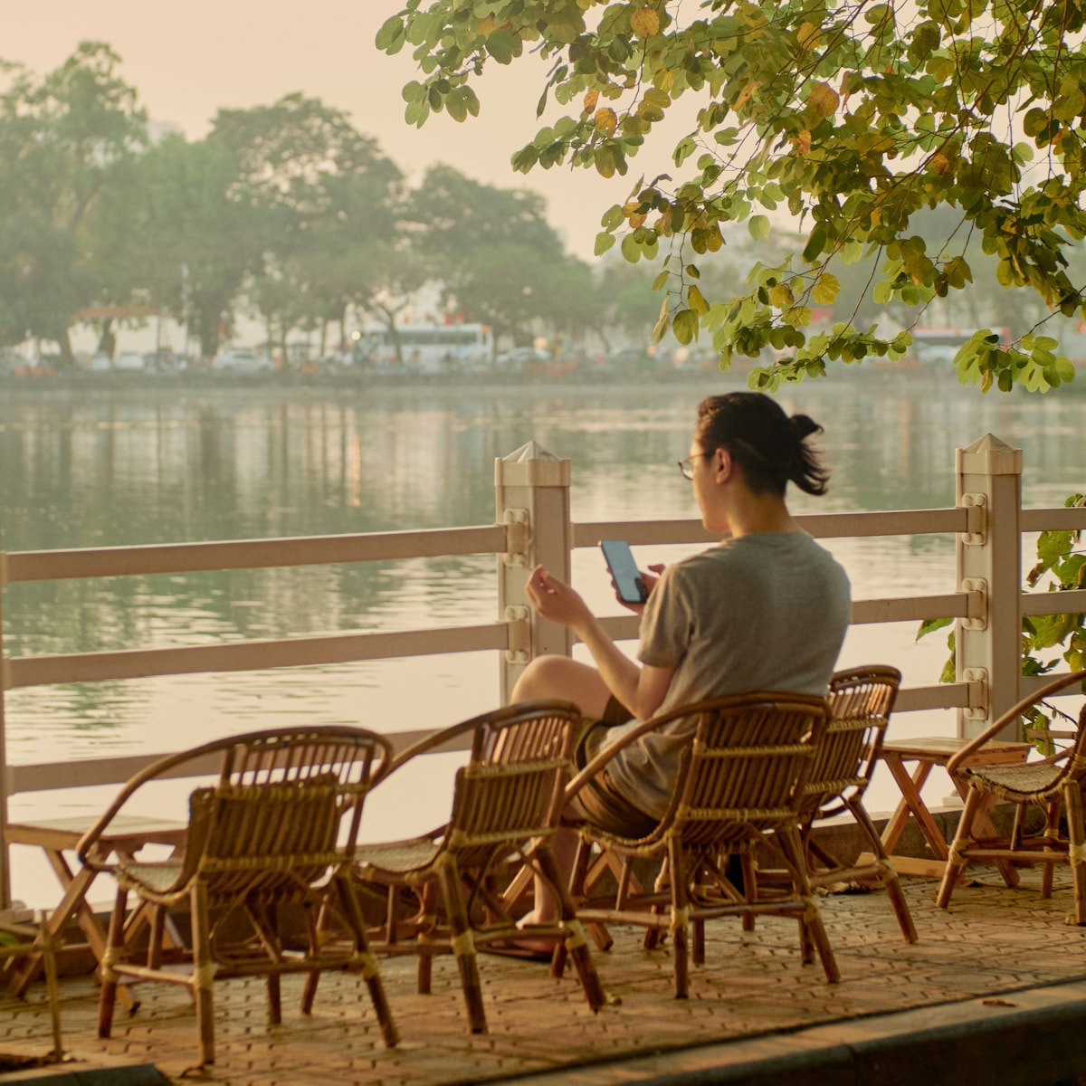 A person is seated at a wooden railing by a serene waterway, surrounded by several wicker chairs. Soft morning light reflects off the calm water, with greenery framing the scene, creating a peaceful atmosphere for relaxation.