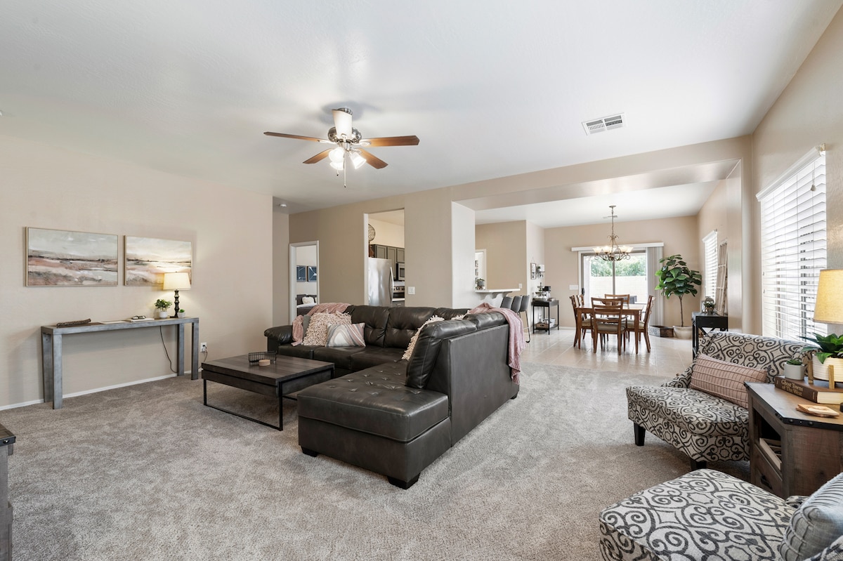 An open living area features a plush sectional sofa, a coffee table, and a ceiling fan. A dining area is visible in the background, accompanied by a well-lit kitchen. Natural light enters through the windows, illuminating the neutral tones and cozy decor.