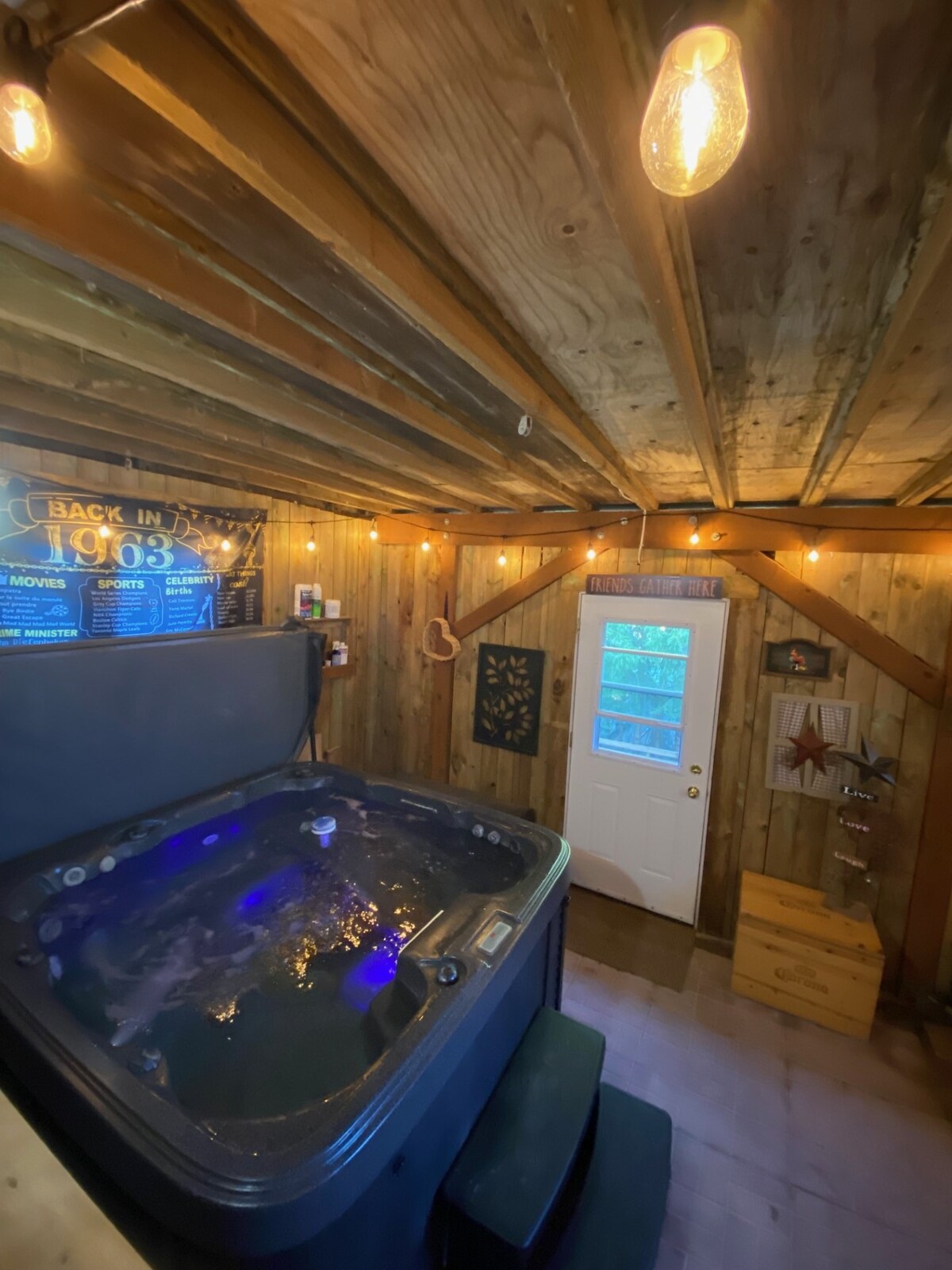 A hot tub is positioned in a cozy room with wooden beams overhead. Soft lighting enhances the relaxing atmosphere, while a door provides access to the outside. The walls are adorned with rustic wood paneling, complemented by a few decorative elements.