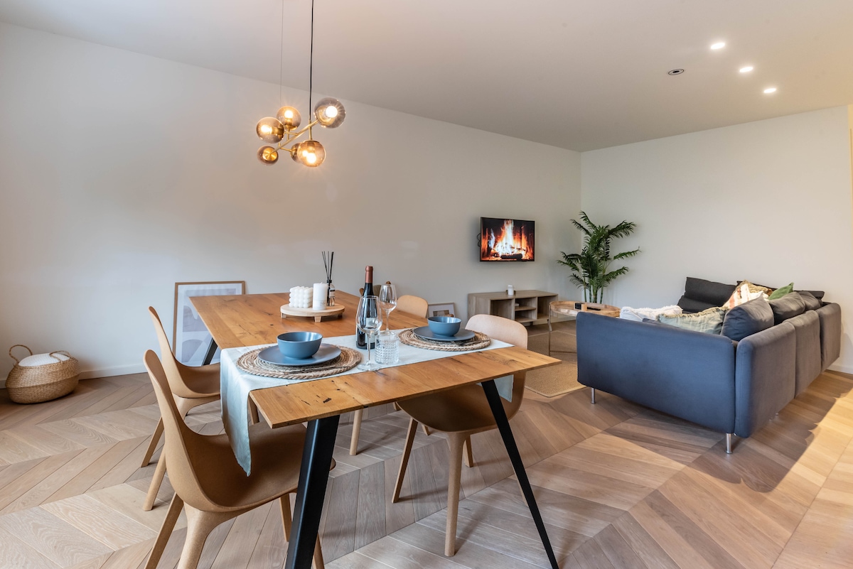 The image displays a modern living area that connects a dining space with a cozy lounge. A wooden dining table is set for meals, accompanied by elegant chairs. In the background, a sofa is positioned near a television, creating a warm and inviting environment.