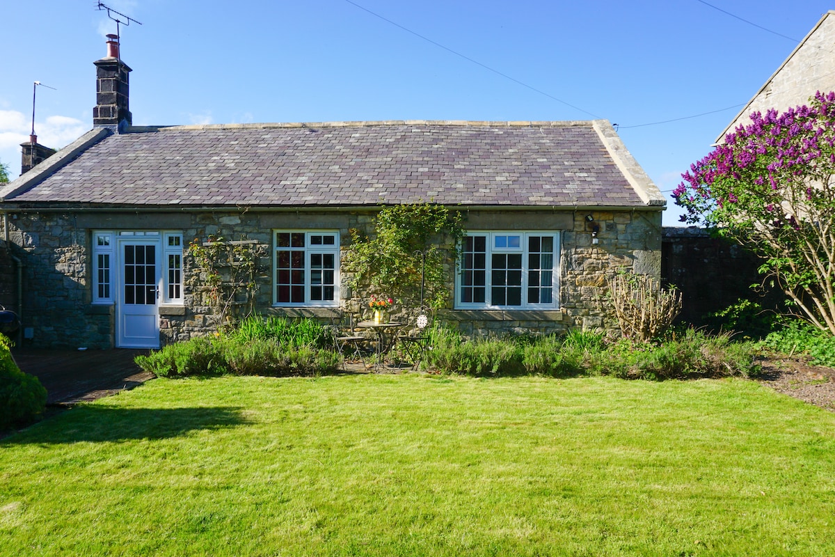 The exterior of a stone cottage is presented with a well-maintained lawn and blooming shrubs. Large windows invite natural light, complementing the inviting entrance. A peaceful atmosphere is suggested by the surrounding greenery and the clear blue sky above.