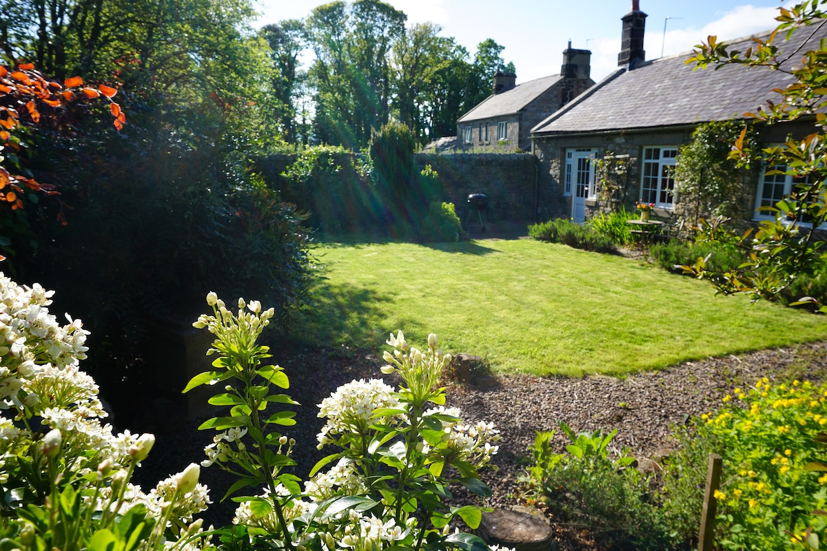 The walled garden is surrounded by greenery, featuring well-maintained grass and blooming flowers. A barbecue grill is positioned on the patio, and the charming cottage exterior can be seen in the background, framed by lush trees and shrubs.