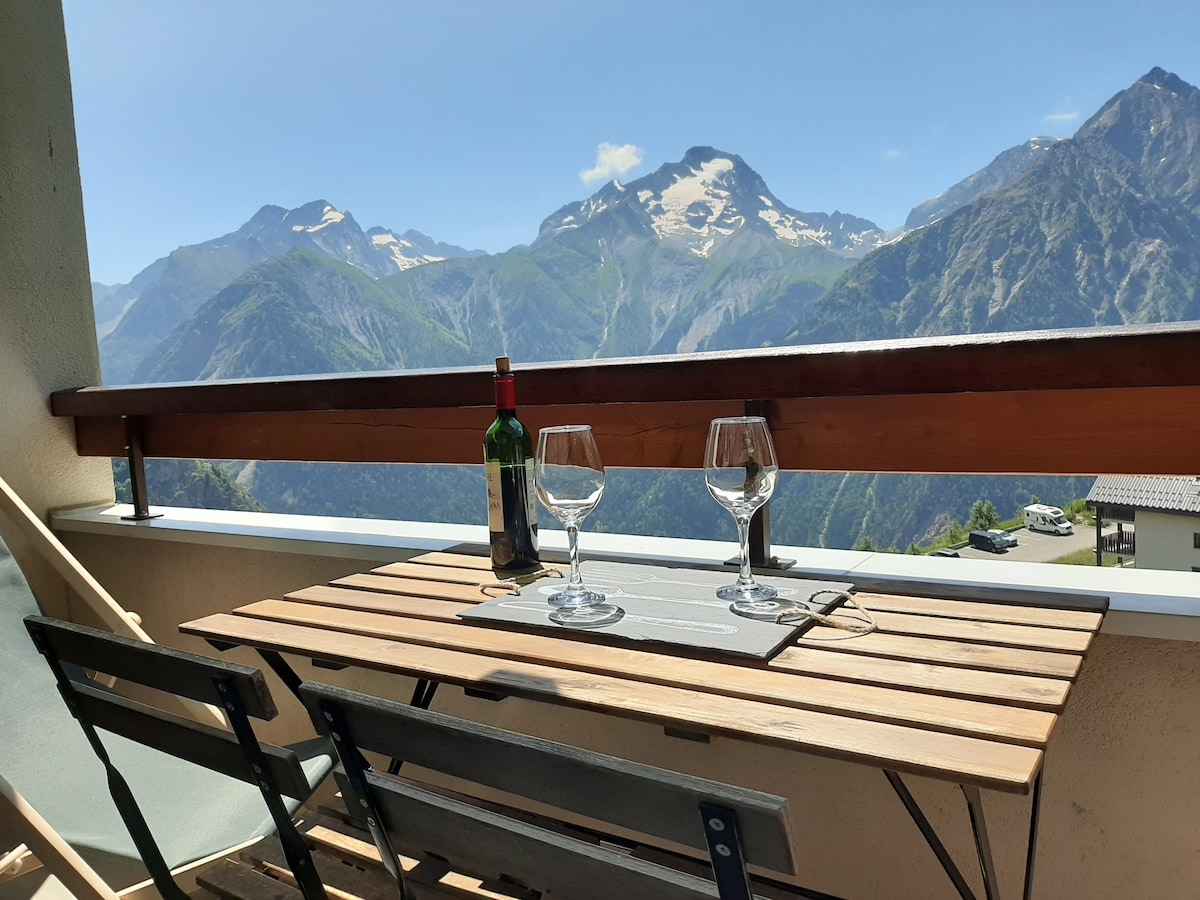 A balcony table is set with two glasses and a bottle of wine, overlooking a stunning mountain view. The table is paired with two wooden chairs, inviting relaxation amid the serene landscape. The bright blue sky complements the scene, enhancing the outdoor experience.