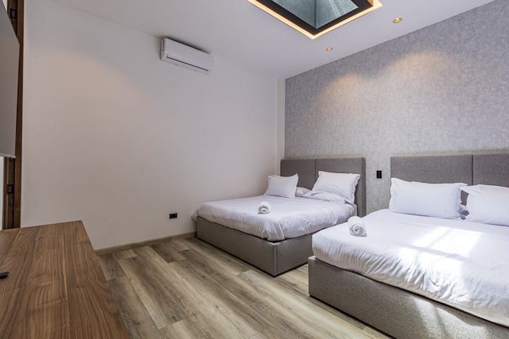 Un dormitorio luminoso y acogedor con dos camas tamaño queen, sábanas suaves, aire acondicionado y un tragaluz que aporta luz natural serena, perfecto para una estancia tranquila y elegante.