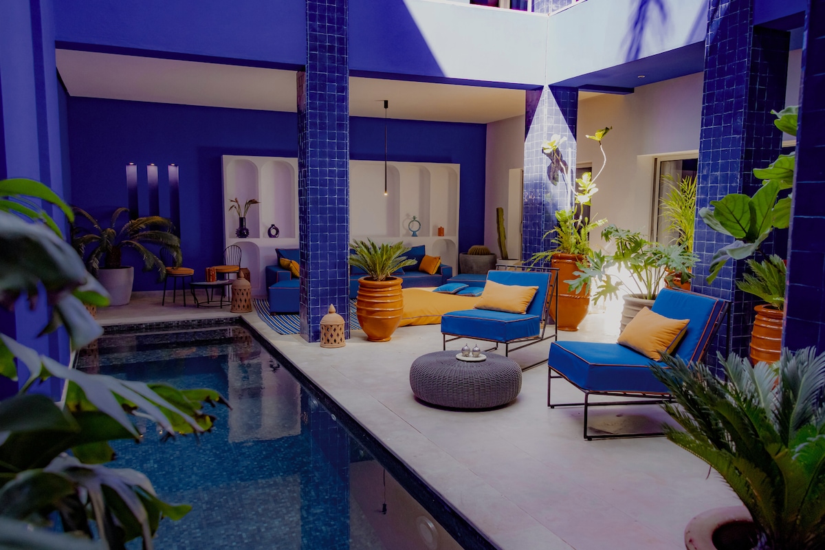 A vibrant courtyard is highlighted by a swimming pool surrounded by blue lounge chairs and decorative plants. Sunlight filters through the open space, illuminating terracotta pots and modern furnishings. The setting offers a tranquil atmosphere for relaxation and socializing.