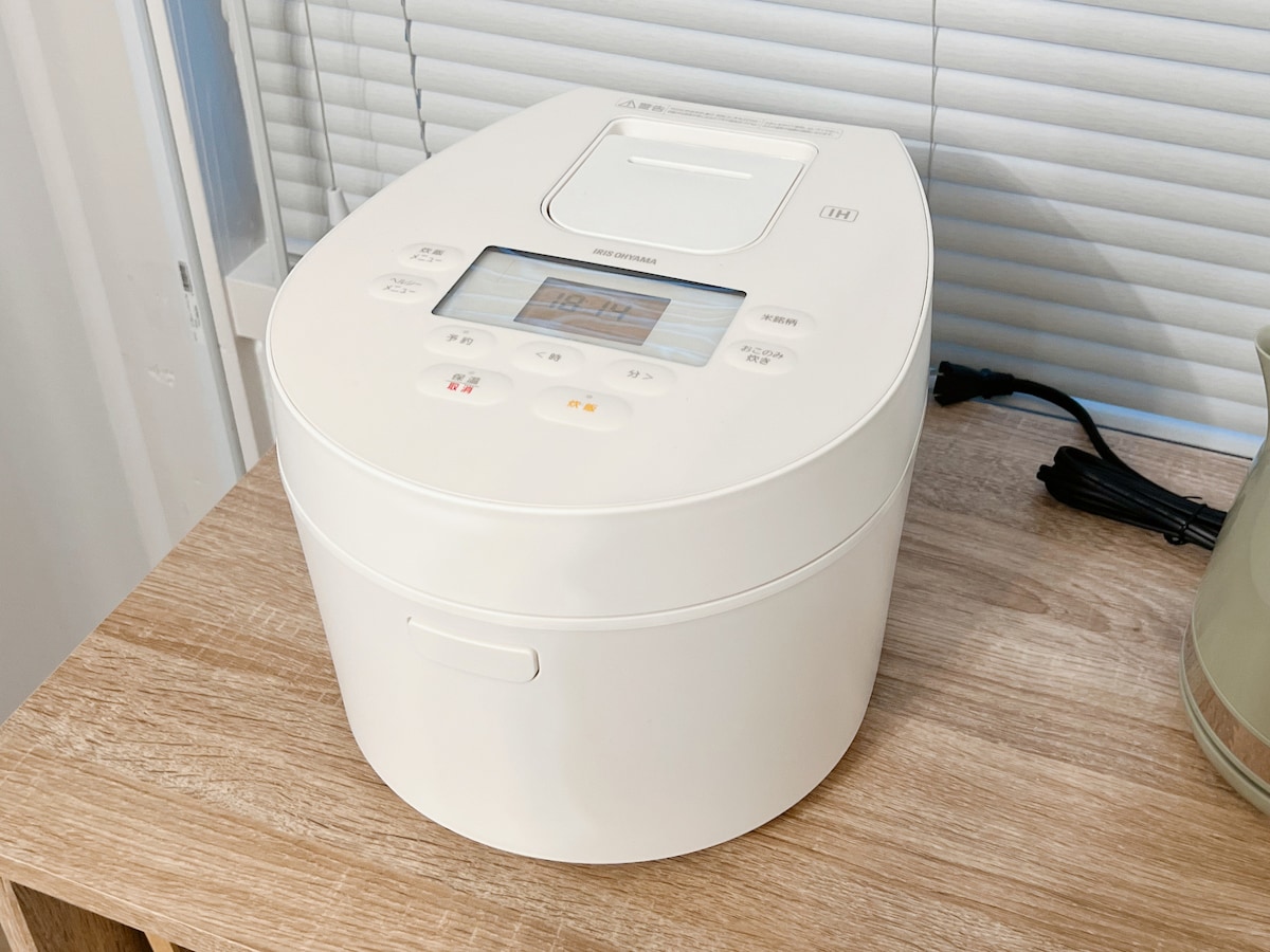 A modern rice cooker sits on a wooden surface, featuring a digital display on the top. The cooker is white, with various buttons and indicator lights clearly visible. Light filtering through nearby blinds highlights the appliance's sleek design.