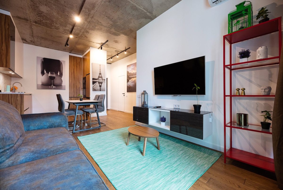 An open living area features a comfortable sofa and a low coffee table on a textured rug. A flat-screen TV is mounted on the wall opposite a dining table with chairs. Bright artwork and a red shelving unit with plants add to the modern aesthetic.