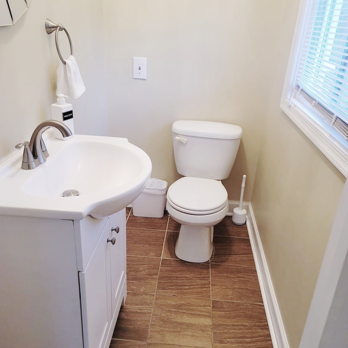 A clean bathroom is shown with a compact sink and a toilet. The sink features a sleek faucet, and a towel is neatly hung nearby. A small window allows natural light to enter, illuminating the neutral-toned walls and tile flooring.
