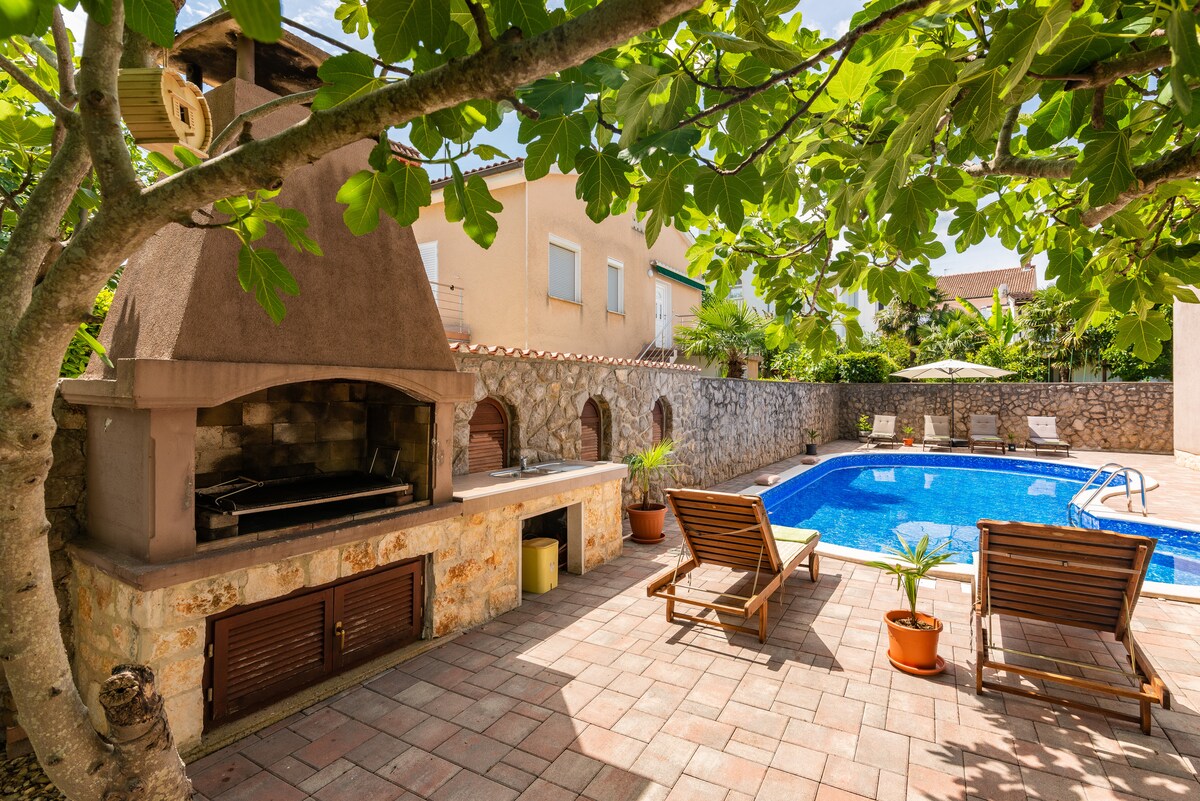 An outdoor area features a charcoal grill beneath a shaded structure, surrounded by stone walls. Two lounge chairs are positioned beside a private pool, which reflects the sunlight. Potted plants add greenery, while a bright umbrella can be seen in the background.