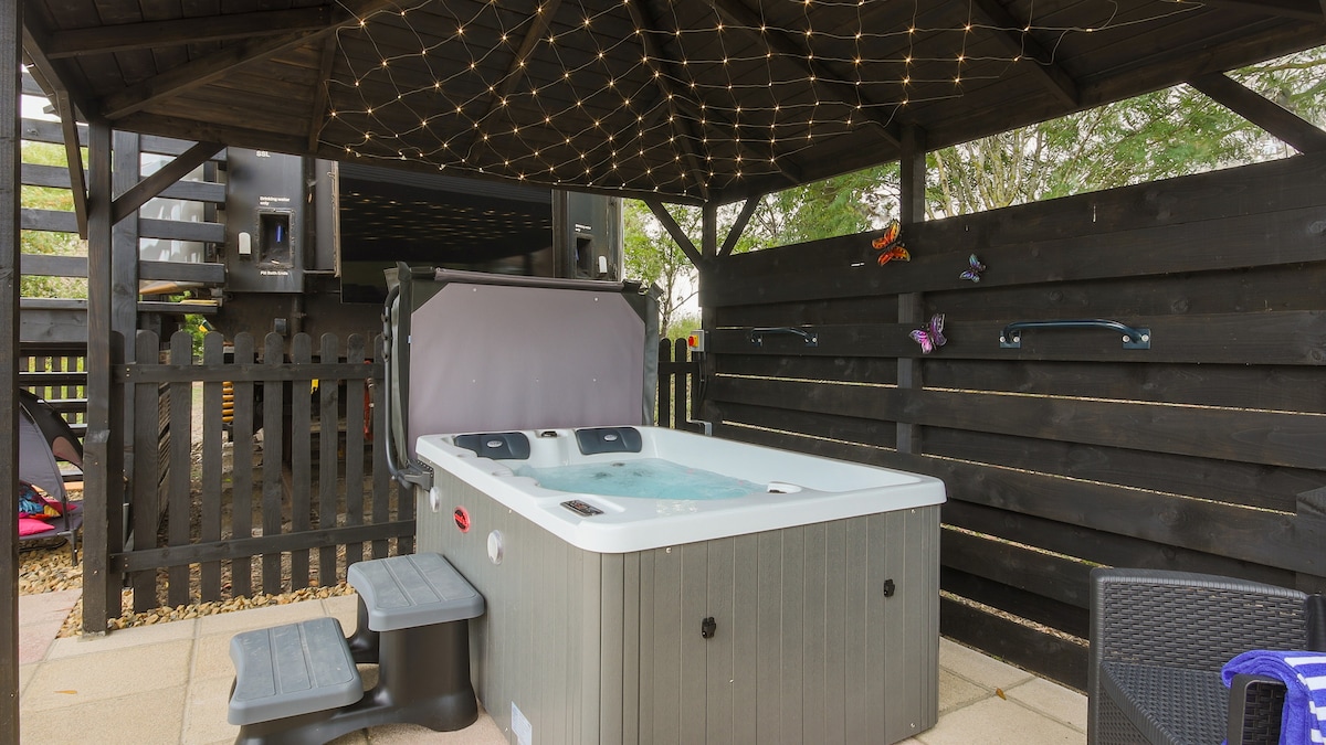 A sheltered hot tub is located within an enclosed garden area, complemented by dim lighting suspended from a wooden gazebo. Surrounding black fencing provides privacy, while a paved surface offers an accessible foundation. Nearby, two steps lead to the tub, ensuring ease of entry.