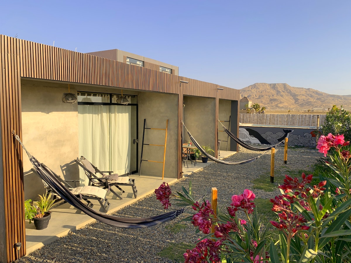 A series of individual accommodation units feature shaded balconies with hammocks and seating areas. Lush tropical plants are positioned in the foreground, while mountains rise in the background under a clear blue sky. Natural materials and textures contribute to a relaxed atmosphere.