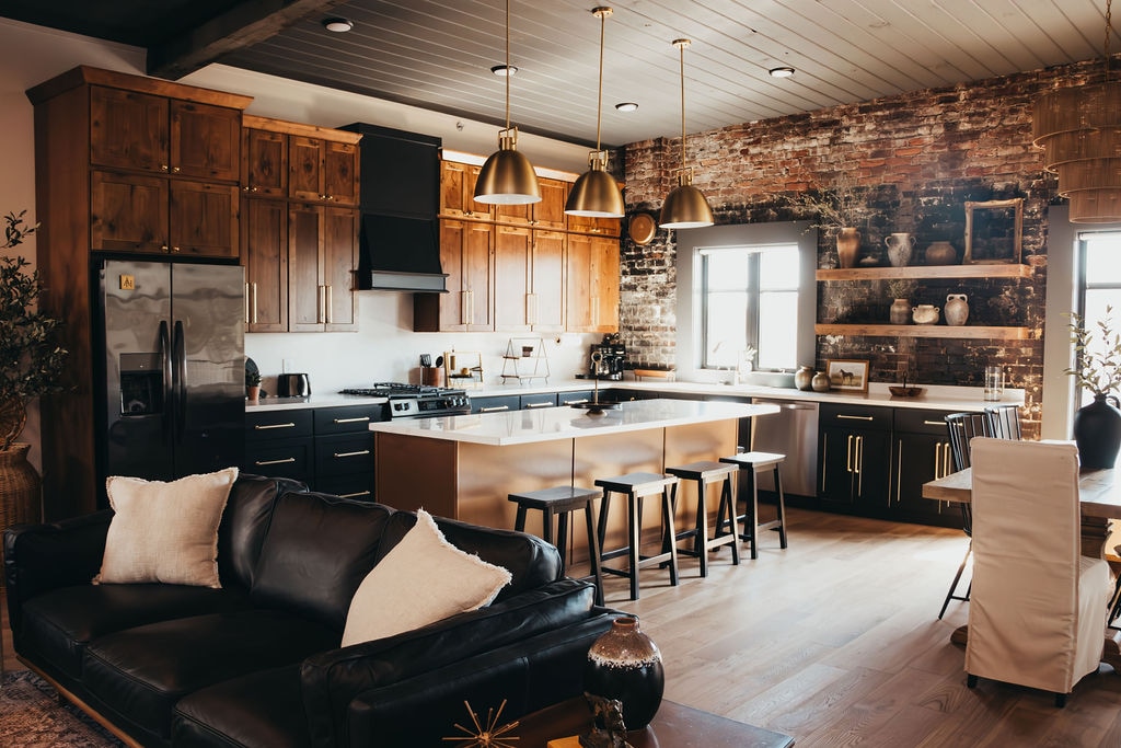 The kitchen features modern stainless steel appliances and a large central island with seating for four. Rich wooden cabinetry complements the exposed brick wall, while warm pendant lights illuminate the space. A cozy living area is nearby, featuring a dark leather sofa and decorative accents.