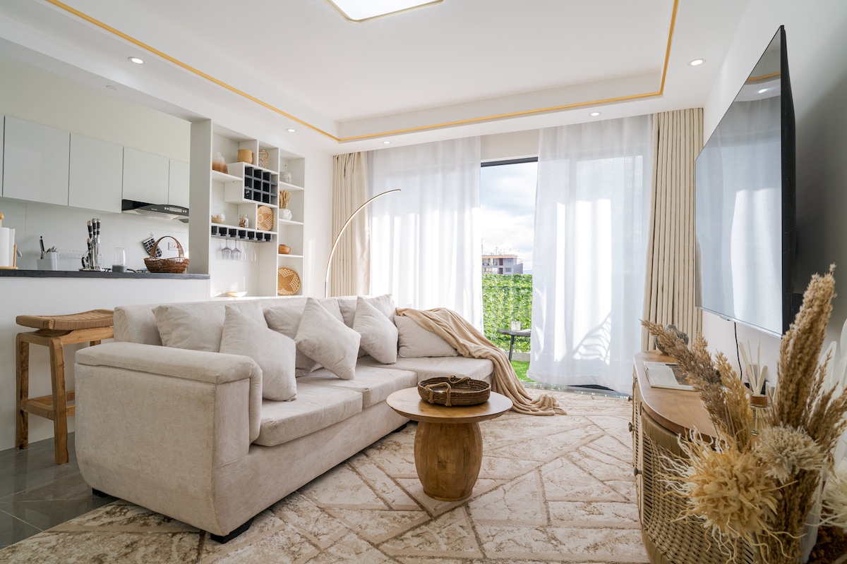 A spacious living area is highlighted by a soft beige sectional sofa and a round wooden coffee table. Natural light fills the space through large glass doors that lead to a private balcony. Light, airy curtains frame the seating area, creating a calm ambiance.