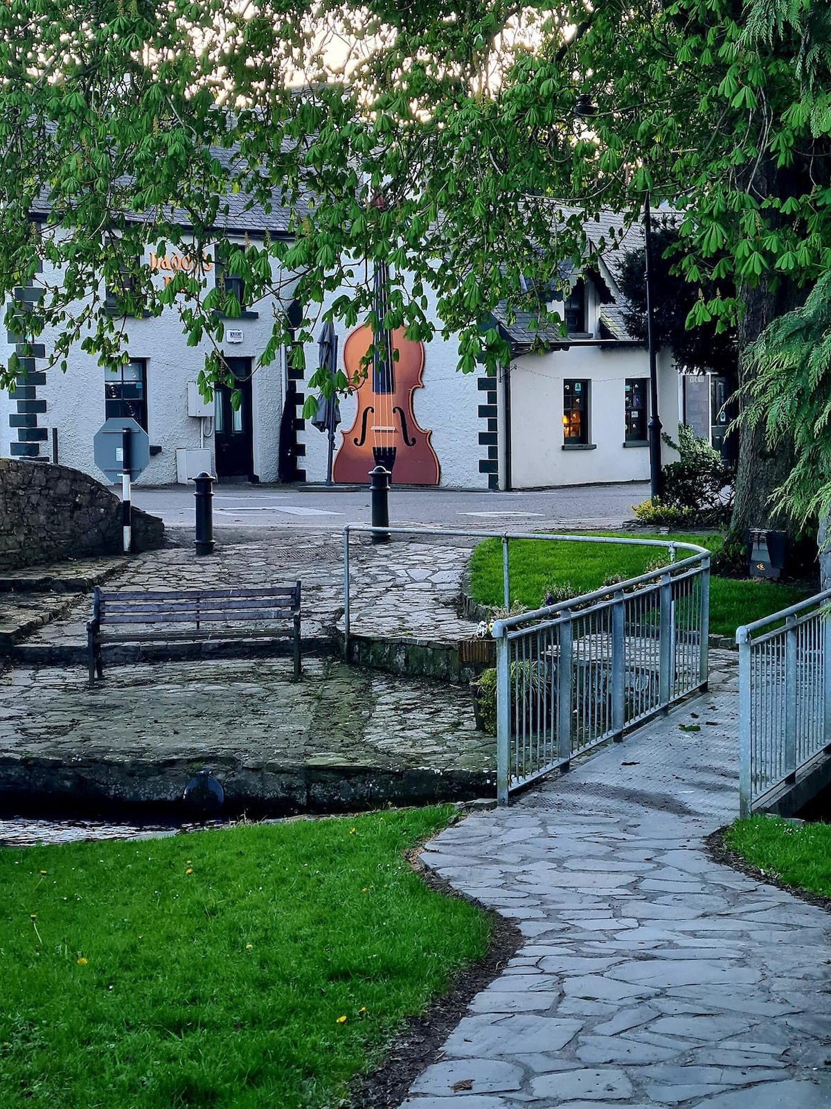 A stone pathway winds through a grassy area leading towards a bridge. In the background, a white building with a large painted violin decorates its wall, accompanied by softly lit windows. Trees provide greenery around the scene, enhancing the natural setting.