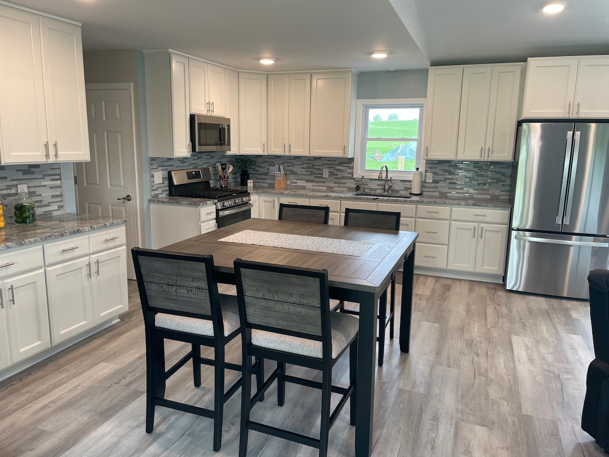A modern kitchen is presented with white cabinetry and stainless steel appliances. A central dining table with six dark chairs is positioned in the foreground. Light-colored countertops complement the grey-toned backsplash, while a large window offers a view of the greenery outside.