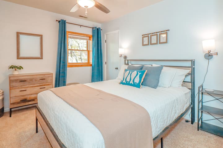 A queen bed with closet, dresser, tv, end table, ceiling fan, and a bathroom next door. The window has light filtering mini blinds and room darkening curtains.