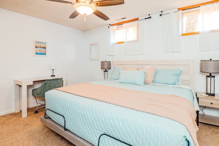 A king bed with end tables, dresser, closet, tv, ceiling fan, and a dedicated workspace. There is a bathroom next door. The windows have room darkening shades and light filtering curtains.