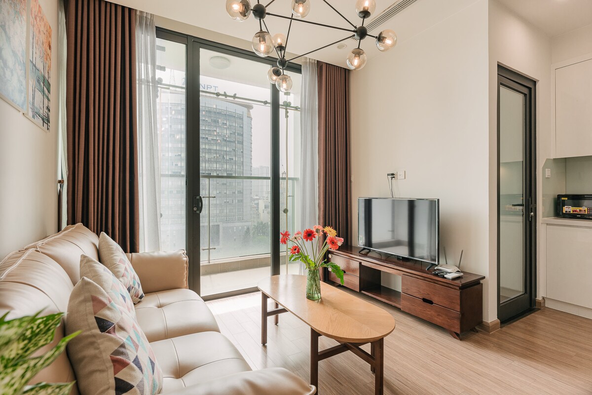 A modern living area features a comfortable sofa with decorative cushions, a wooden coffee table, and a flat-screen TV. Large glass doors open to a balcony, allowing natural light to fill the space. A vase of fresh flowers adds a touch of color.