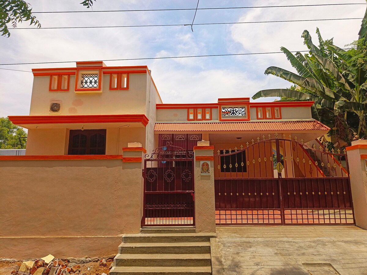 A charming two-story house featuring a welcoming facade with orange accents. The gated entrance provides privacy, while lush greenery surrounds the property. A smooth pathway leads to the entrance, enhancing the overall inviting appearance of the home.