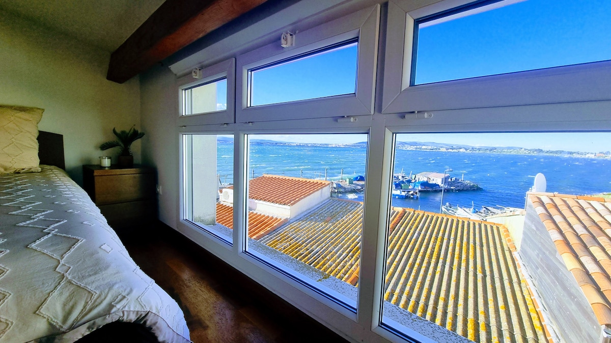 A serene view is seen from large windows revealing the expansive étang. Bright blue waters are dotted with boats while the sun illuminates the nearby rooftops. The inviting space includes a bed with soft linens, complemented by natural light pouring in from the windows.