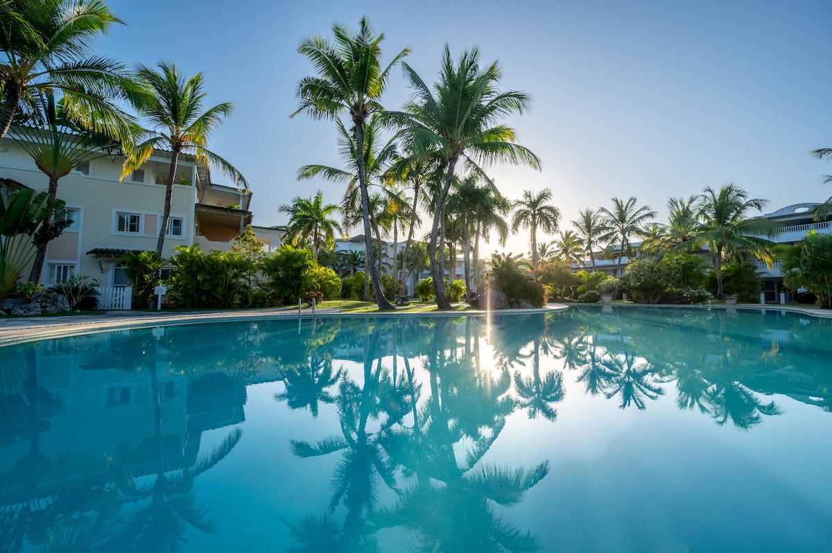 A large swimming pool reflects the surrounding palm trees and vibrant greenery. The sunlight casts a warm glow over the tranquil water, creating a serene atmosphere. The background features well-maintained resort buildings, enhancing the tropical setting.