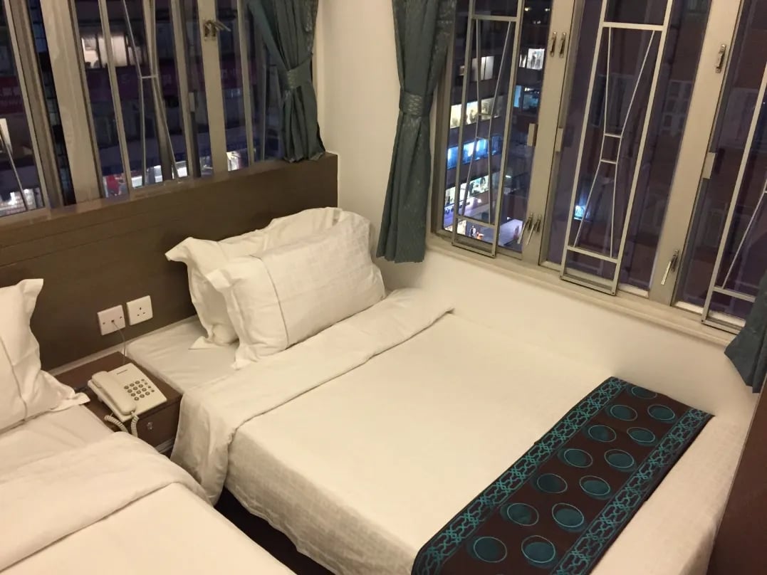 A well-organized guest room displays two beds with white bedding, positioned beside large windows with metal grilles providing views of the city. A telephone rests on the bedside table, and curtains in a subtle blue frame the windows.