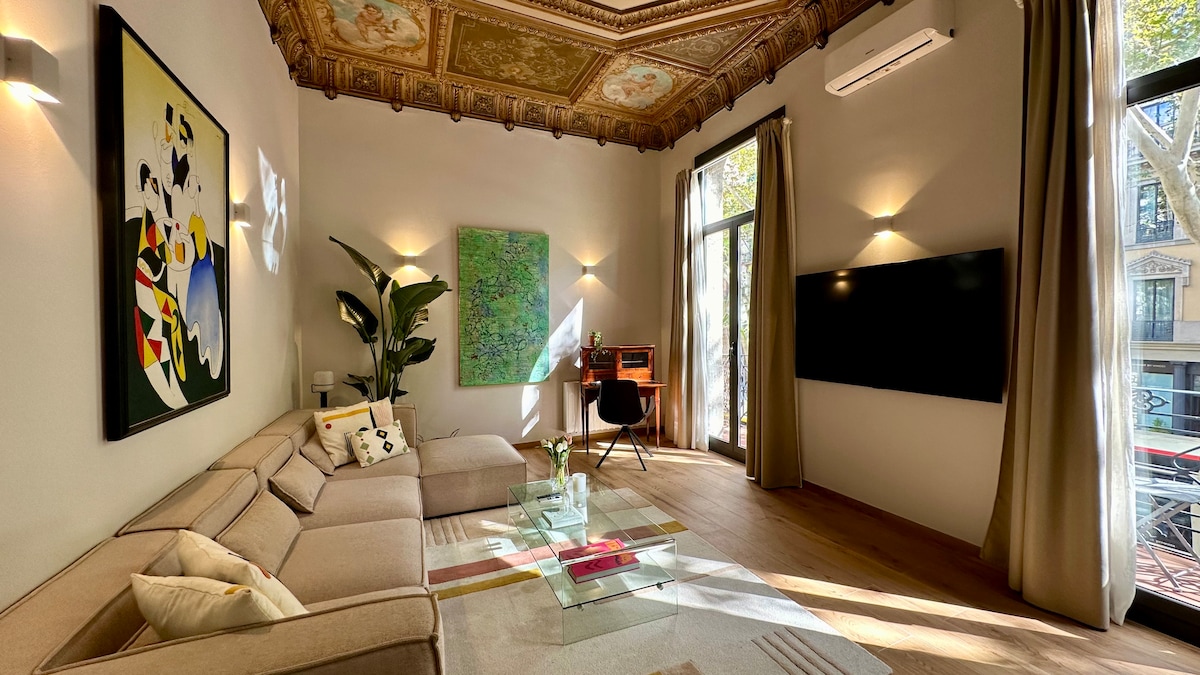 A spacious living room features a large beige sectional sofa accompanied by decorative pillows. Floor-to-ceiling windows allow natural light to illuminate the room, showcasing high, ornately decorated ceilings. An abstract painting adds color to the wall, while a modern television is mounted nearby.