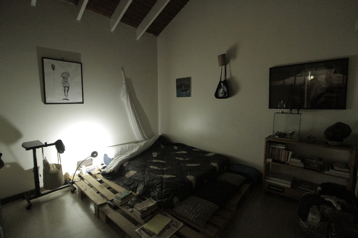 A minimalist bedroom is presented with a low-profile mattress placed on wooden pallets. Soft lighting is provided by a standing lamp beside the bed. An art piece hangs on the wall, and a small TV is mounted opposite the bed. Various cushions are arranged on the floor.