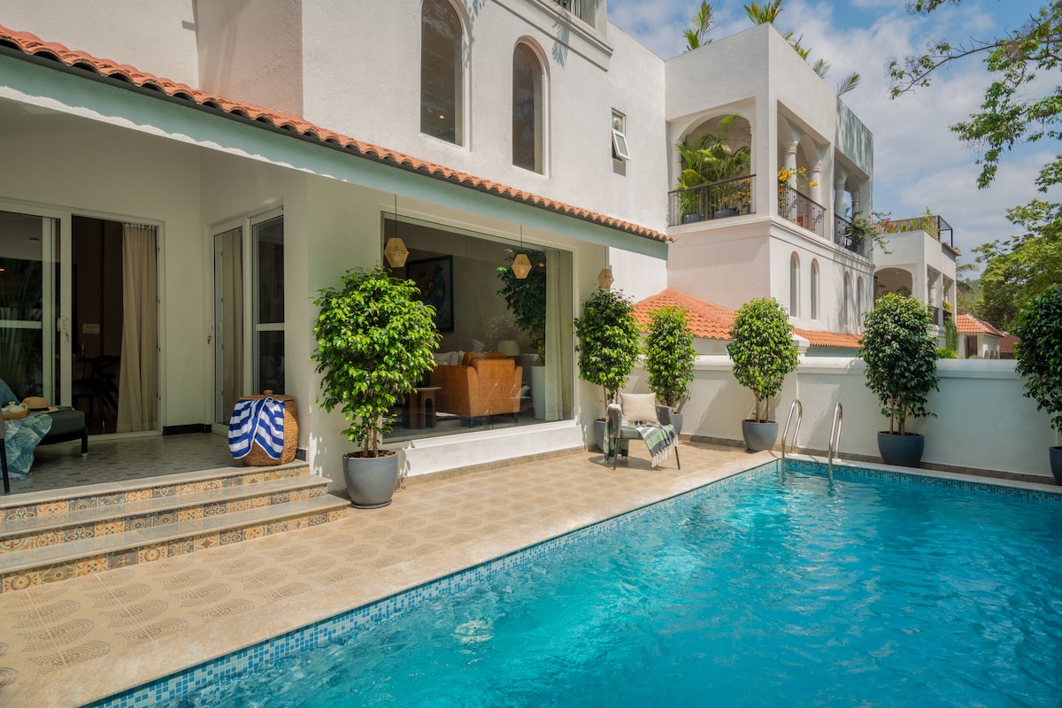 A private pool is framed by lush greenery and paved stone tiles. The villa's expansive glass doors lead to an inviting living area, where natural light enhances the spaciousness. A relaxing outdoor space is complemented by lounge chairs and vibrant potted plants.