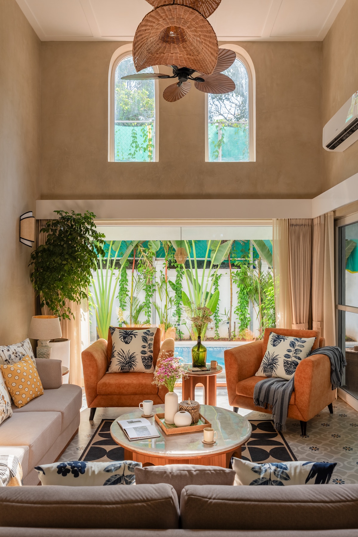 A bright living area is enhanced by high ceilings and large windows, allowing natural light to flood the space. Two inviting orange armchairs are paired with a grey sofa, adorned with decorative pillows. A green plant adds a touch of nature, while a coffee table holds candles and floral arrangements.